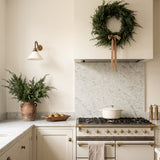 A styled kitchen interior featuring a cream-colored vintage-style gas range with brass handles, a white marble backsplash, a green pine wreath hanging on the range hood, and a potted fern on the counter.