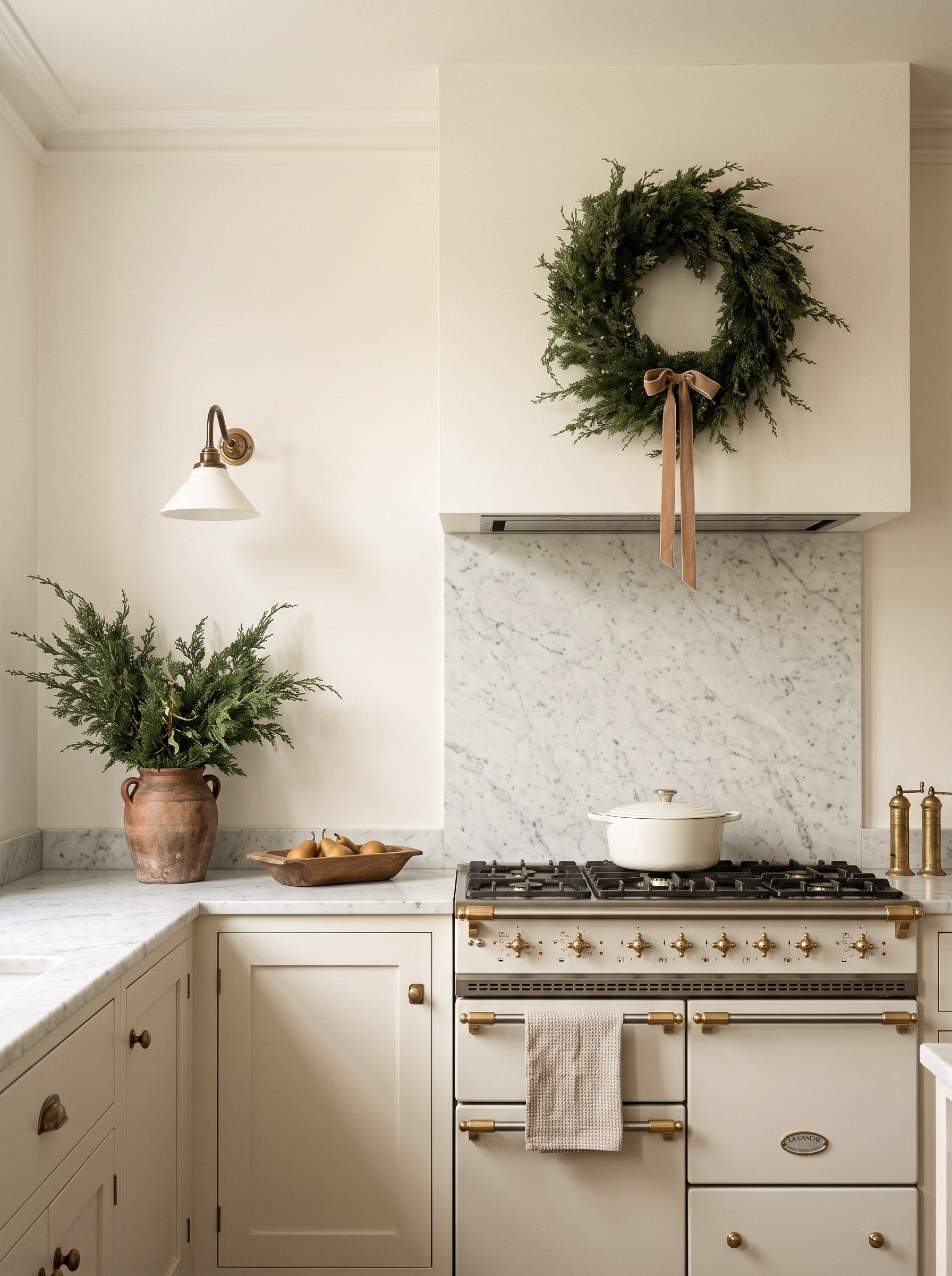 A styled kitchen interior featuring a cream-colored vintage-style gas range with brass handles, a white marble backsplash, a green pine wreath hanging on the range hood, and a potted fern on the counter.