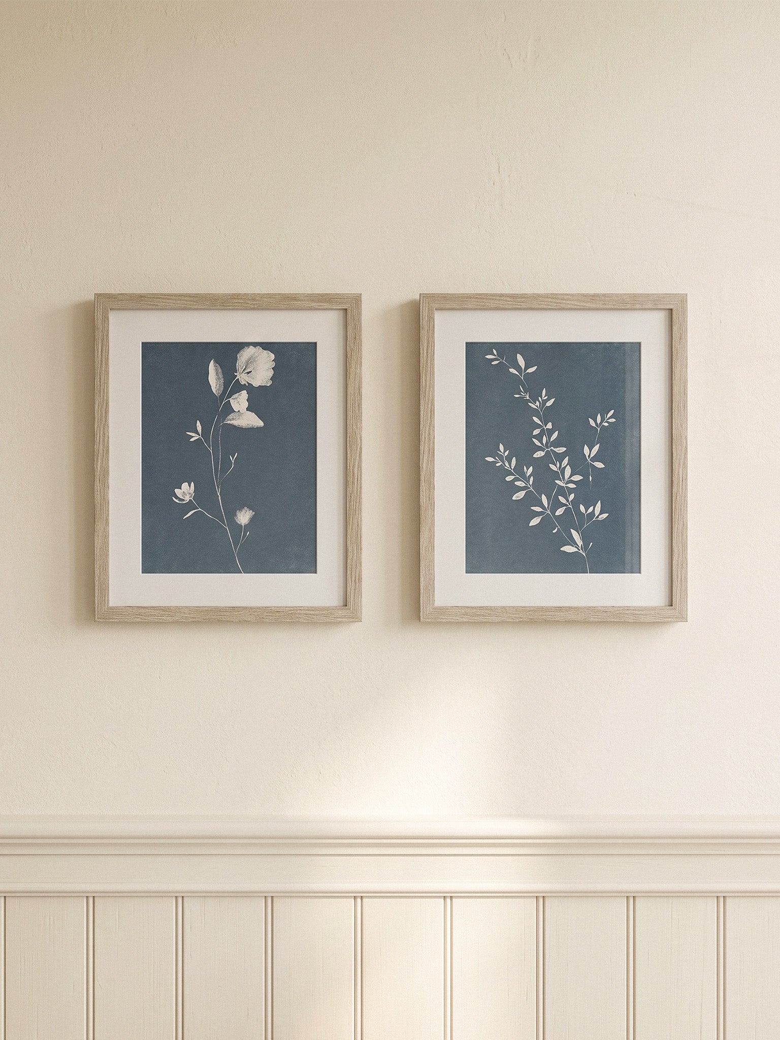 Two framed botanical art prints with blue-gray backgrounds and white floral illustrations, hung side by side on a cream wall above white wainscoting.