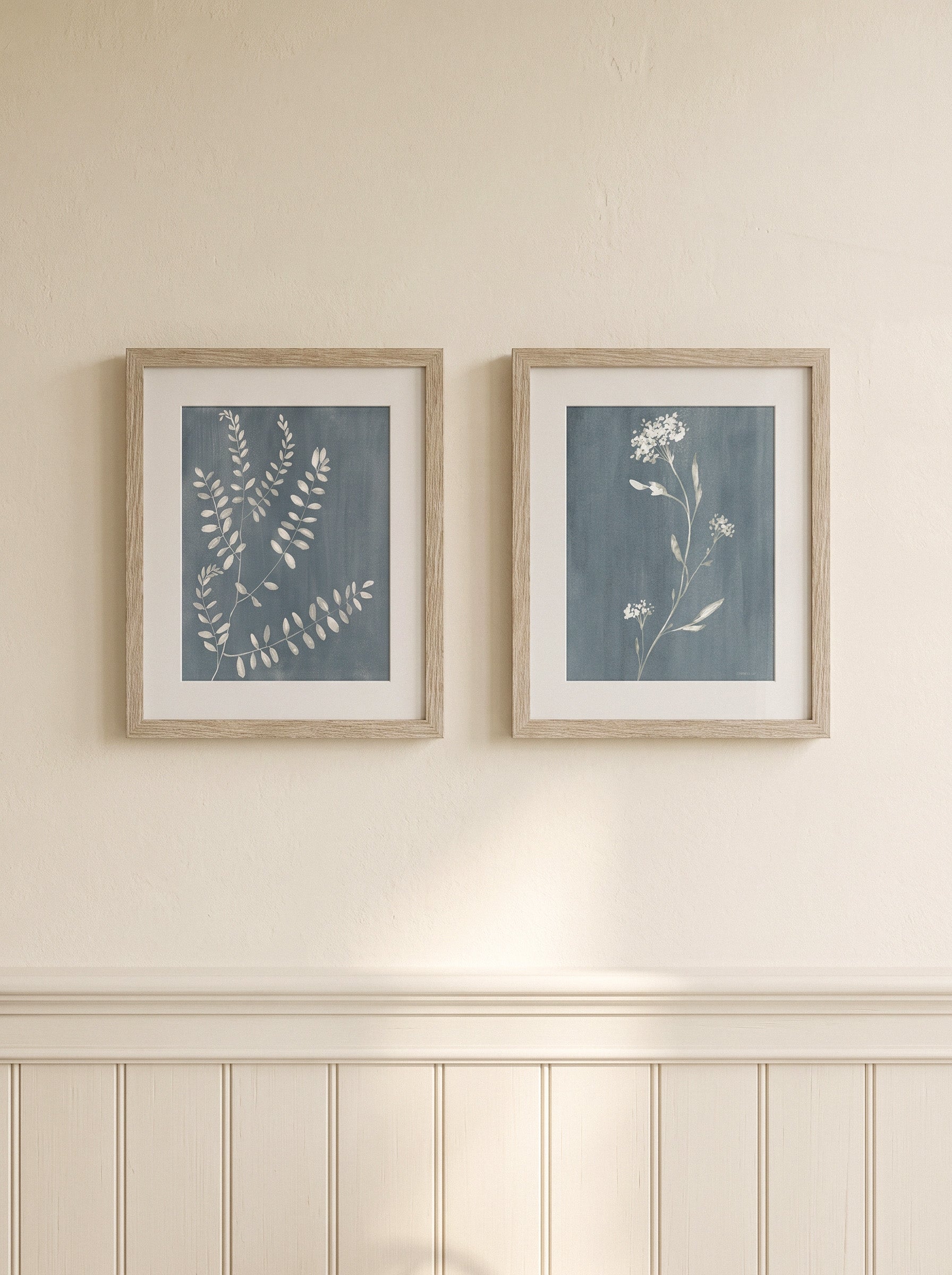 Two framed botanical art prints with blue-gray backgrounds and white floral/leaf illustrations, hung side by side on a cream wall above white wainscoting.