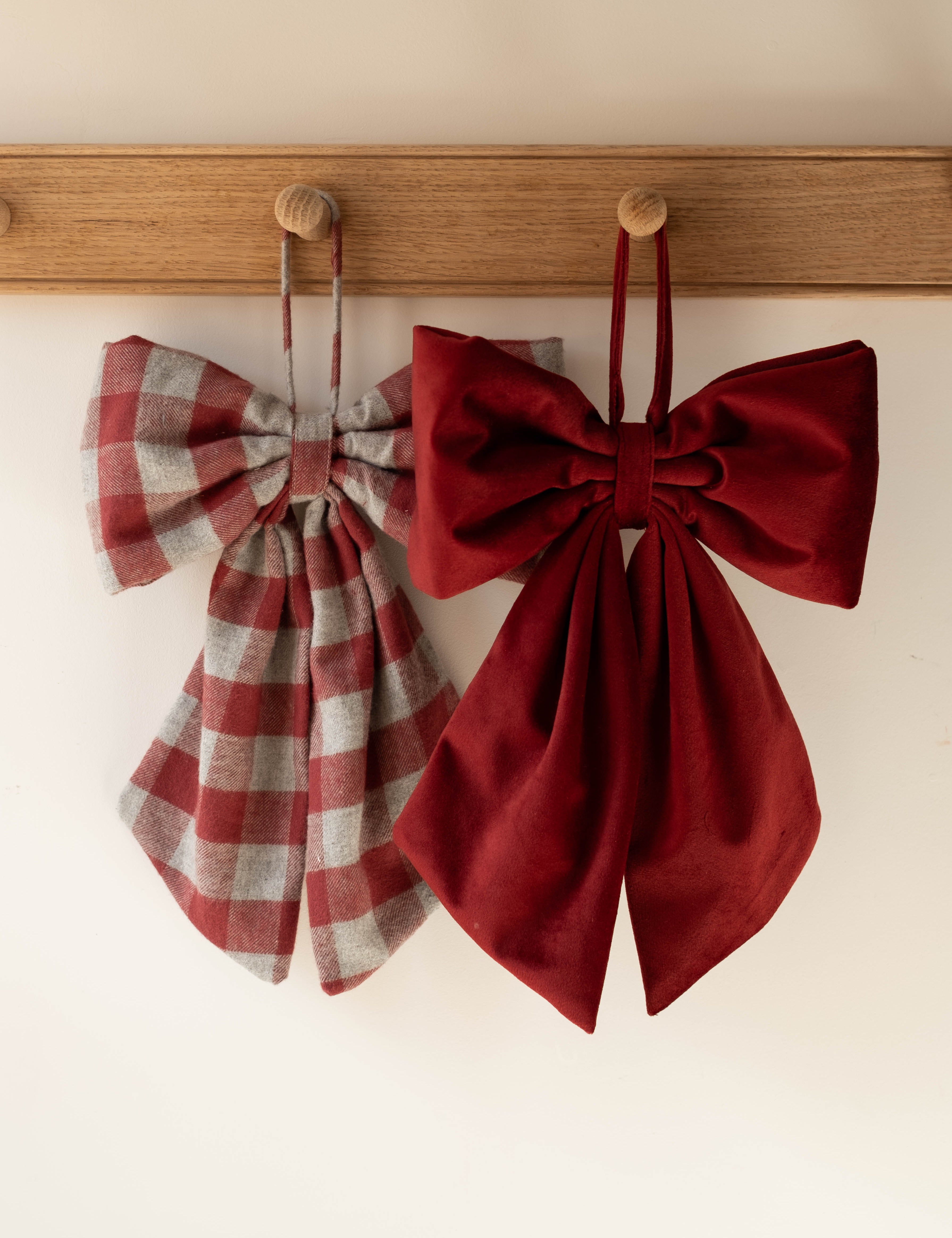 Two fabric bow accessories hanging from wooden pegs on a wooden rail against a plain light wall. The left bow has a muted red and gray plaid pattern, and the right bow is a solid deep burgundy red.