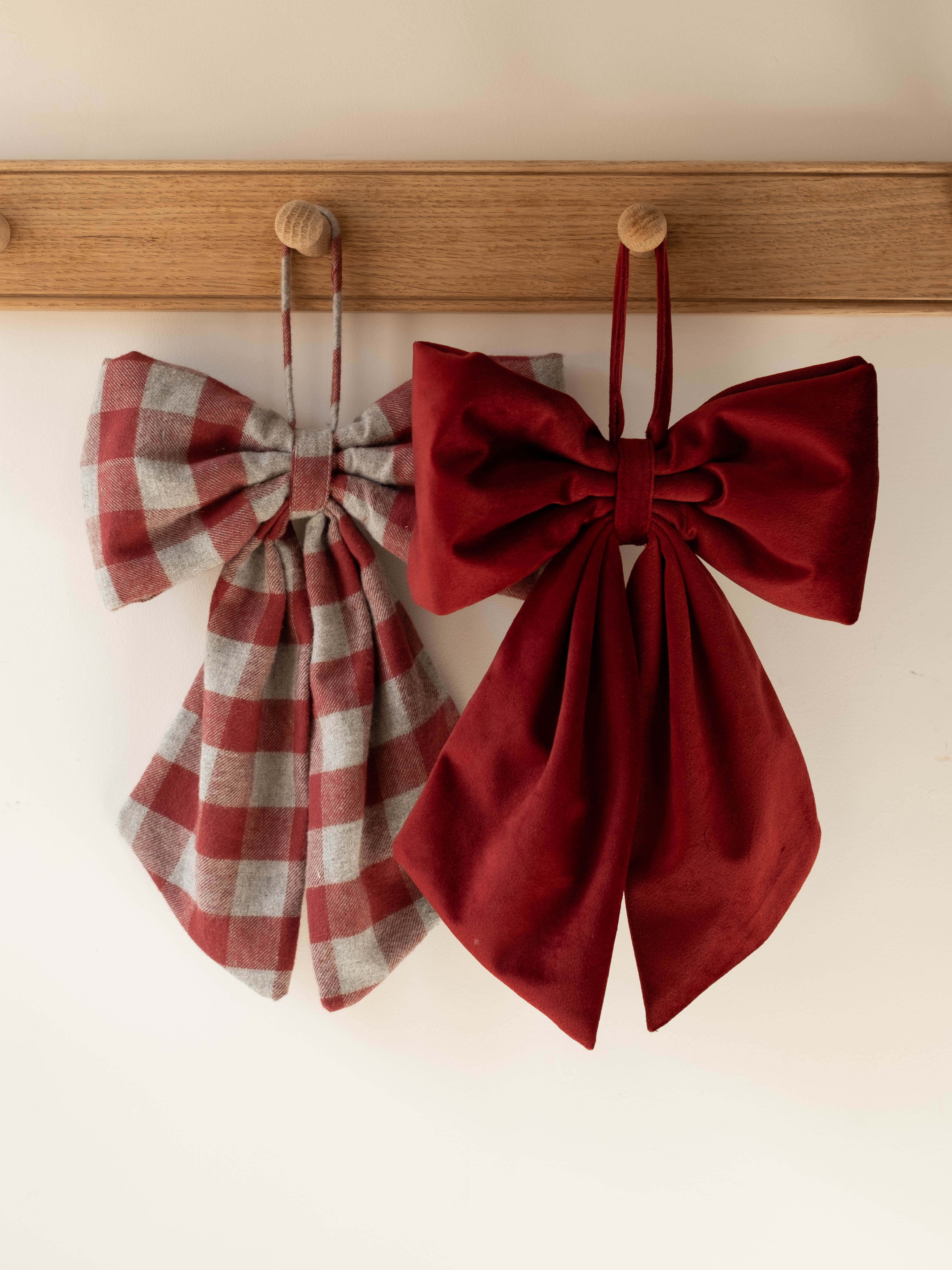Two fabric bow accessories hanging from wooden pegs on a wooden rail against a plain light wall. The left bow has a muted red and gray plaid pattern, and the right bow is a solid deep burgundy red.