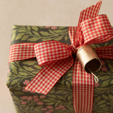A wrapped gift box with olive green and pink floral patterned paper, tied with a red and white gingham ribbon and a small metallic gold bell, on a neutral beige background.