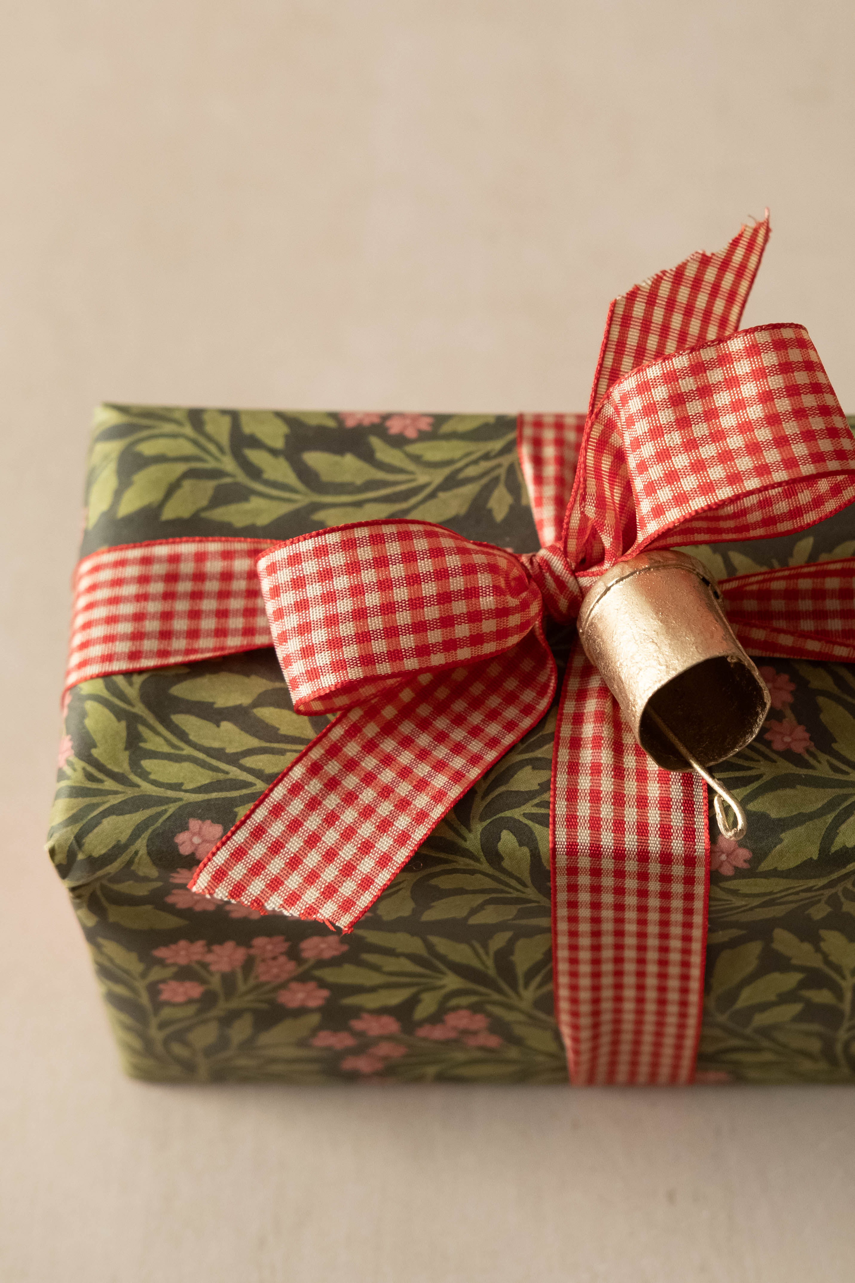 A wrapped gift box with olive green and pink floral patterned paper, tied with a red and white gingham ribbon and a small metallic gold bell, on a neutral beige background.