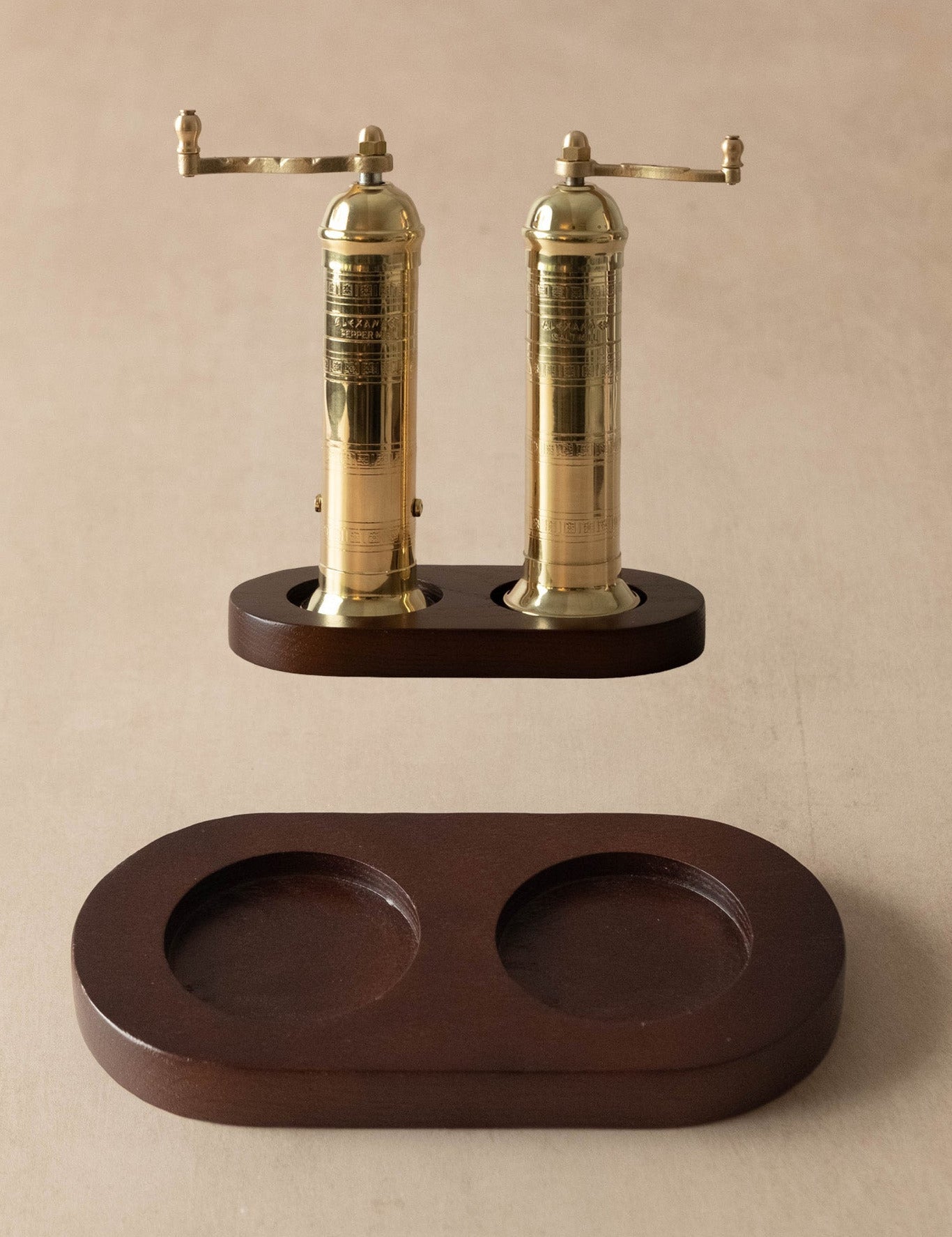 A pair of brass pepper mills with dark wood bases, shown with their matching dark wood storage tray on a neutral beige background.