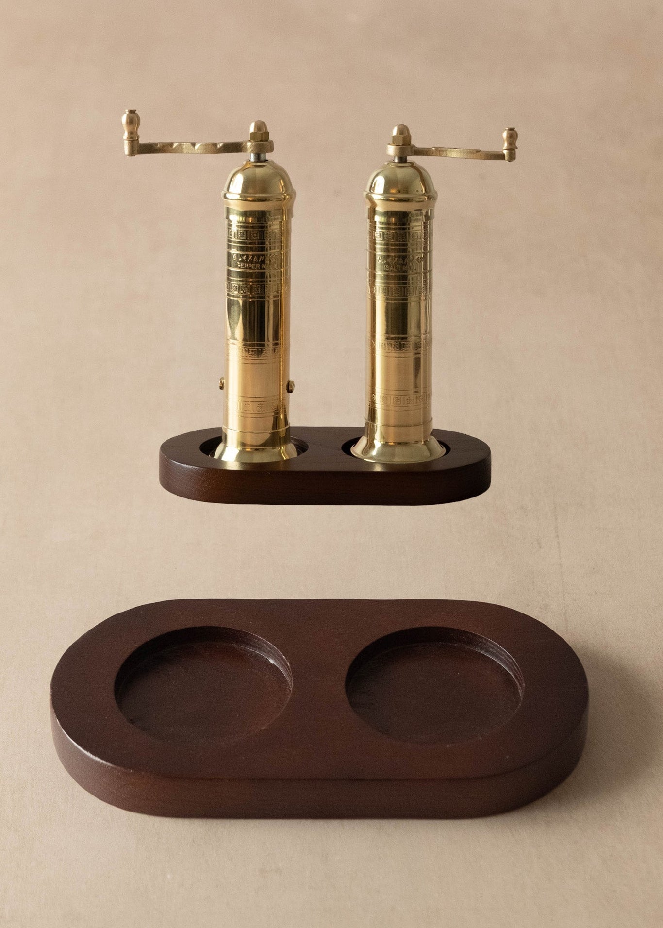 A pair of brass pepper mills with dark wood bases, shown with their matching dark wood storage tray on a neutral beige background.