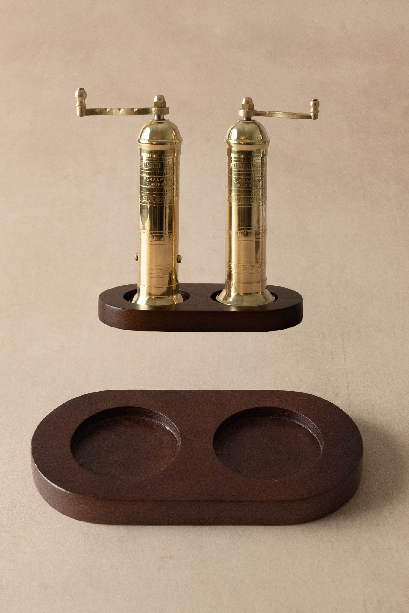 A pair of brass pepper mills with dark wood bases, shown with their matching dark wood storage tray on a neutral beige background.