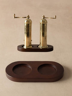 A pair of brass pepper mills with dark wood bases, shown with their matching dark wood storage tray on a neutral beige background.