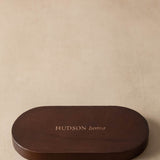 A dark brown oval wooden box lid with the brand name 'HUDSON home' printed in gold lettering, set against a textured beige background.