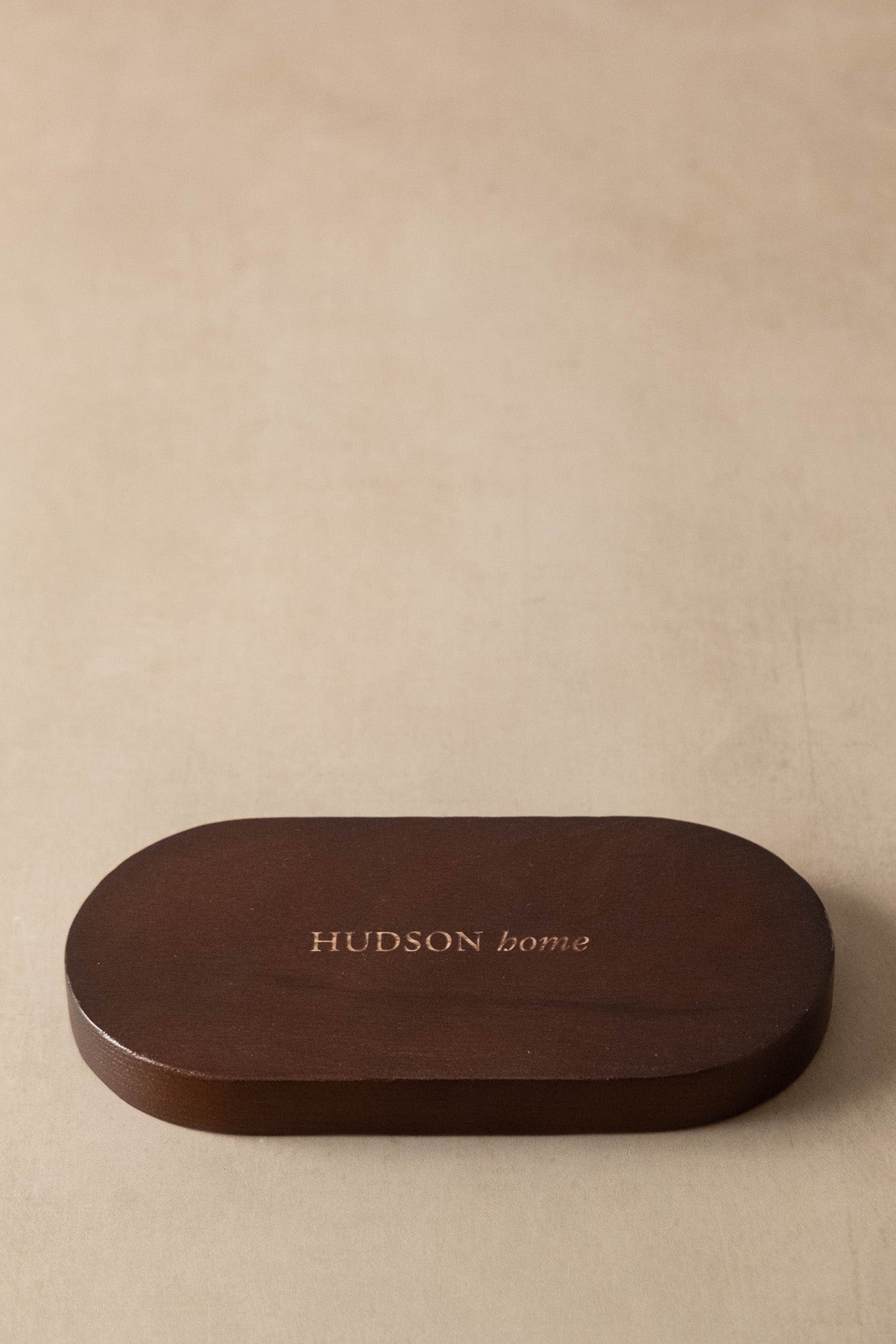 A dark brown oval wooden box lid with the brand name 'HUDSON home' printed in gold lettering, set against a textured beige background.